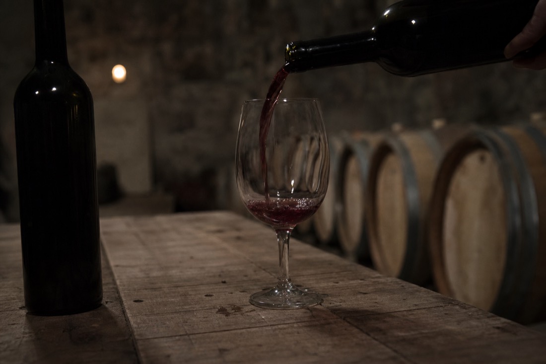 Dry red wine poured into a glass on a rustic table beside an unlabeled dark bottle in cellar light
