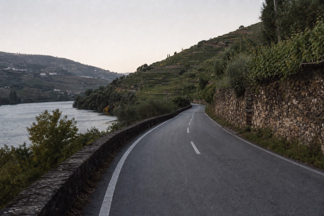 Curve of the N222 road hugging the Douro river with schist walls and terraced vineyards rising on both sides