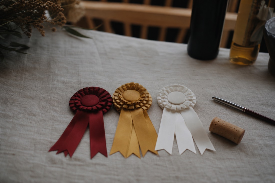Three unlabeled ribbon rosettes resting on a linen cloth beside a wine cork and a pen