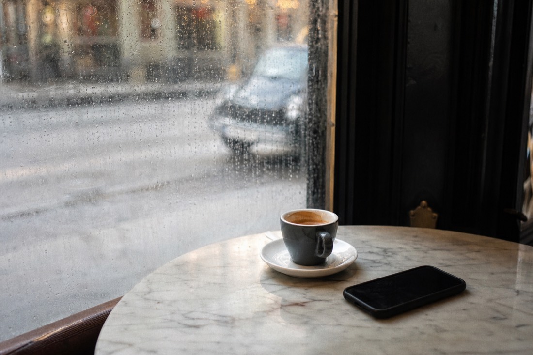 Espresso cup and phone on a marble cafe table beside a rain-speckled window, an unhurried booking moment