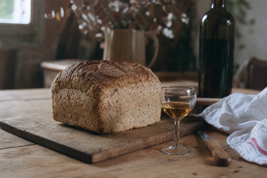 Rustic four-cornered loaf of bread on a wooden board beside a small glass of golden fortified wine