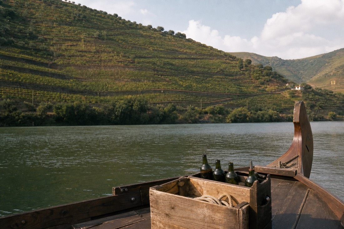 View from a rabelo boat deck over calm river water toward terraced vineyards, wooden bow rising at the frame edge