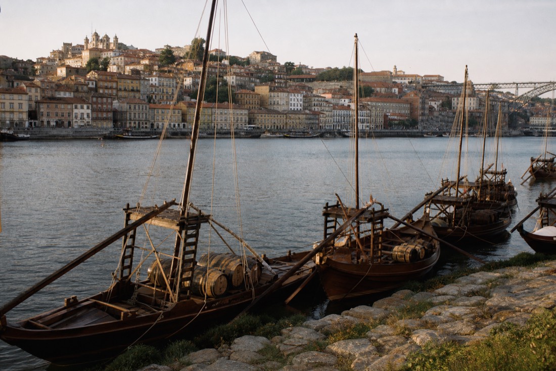 Several moored rabelo boats loaded with display barrels on the riverfront with old town buildings across the water