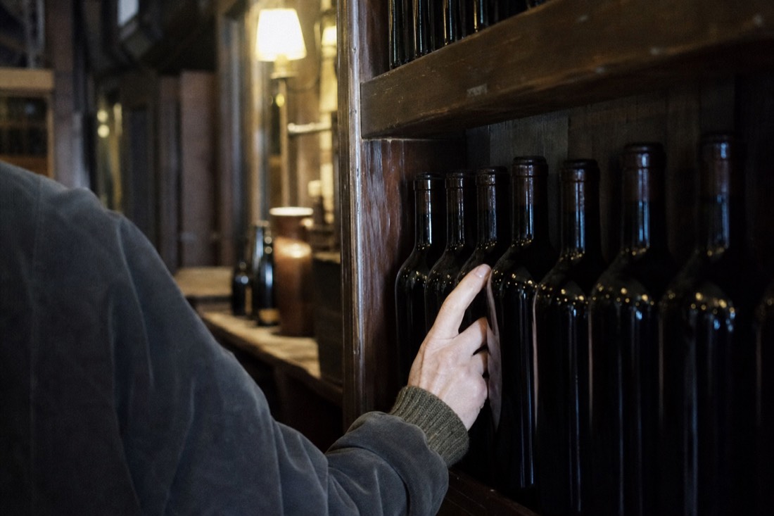 Hand reaching for an unlabeled bottle on a wooden shelf of port bottles in a dim cellar shop