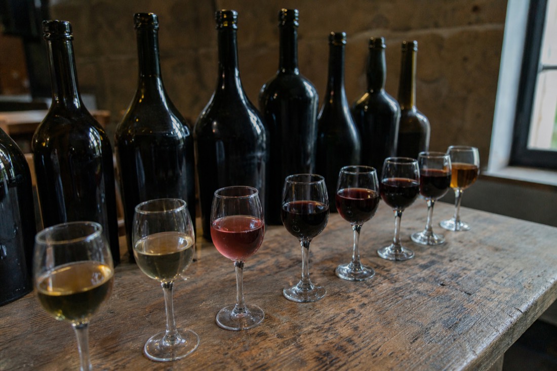 Line of unlabeled port bottles and small tasting glasses showing colors from pale gold through pink to deep ruby and amber
