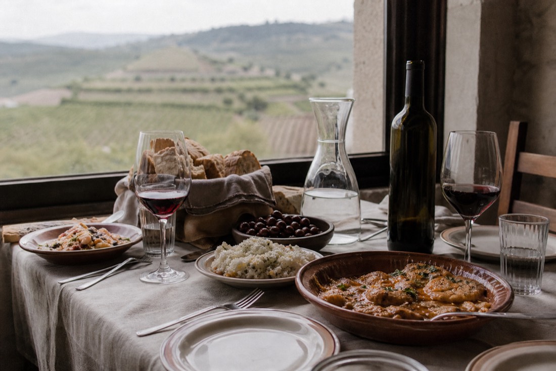 Rustic Portuguese lunch table with regional dishes, bread, olives and glasses of red wine near a window overlooking vineyards
