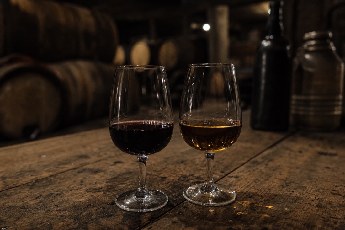 Two glasses of port wine, one garnet and one amber, on a wooden table in soft cellar light