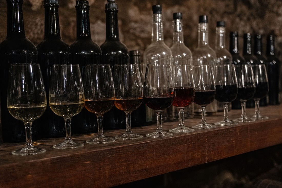 Row of unlabeled port bottles and small tasting glasses showing colors from pale gold to deep ruby