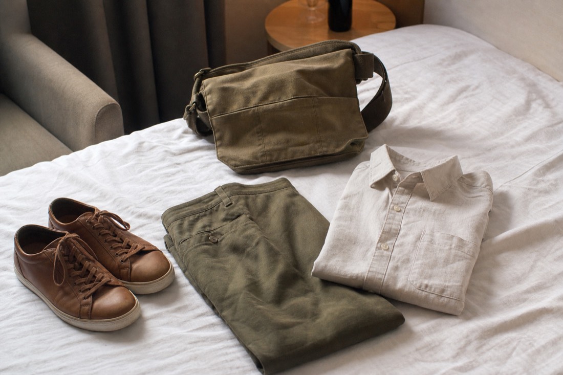 Folded linen trousers, a light shirt and flat leather shoes laid out on a bed beside a small day bag