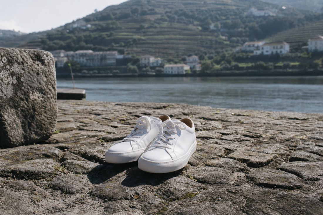 Pair of white leather sneakers standing on worn cobblestones of a riverside quay in a Douro village