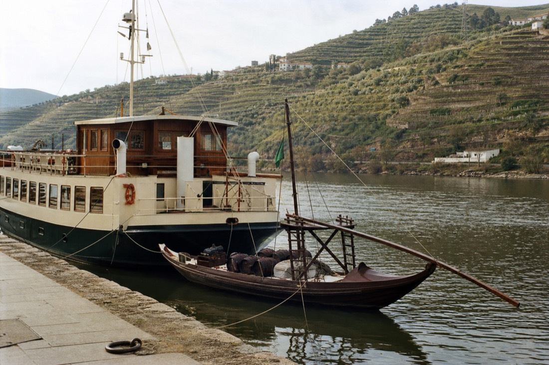 A small wooden rabelo and a larger river vessel moored together at a Douro quay below terraced vineyards