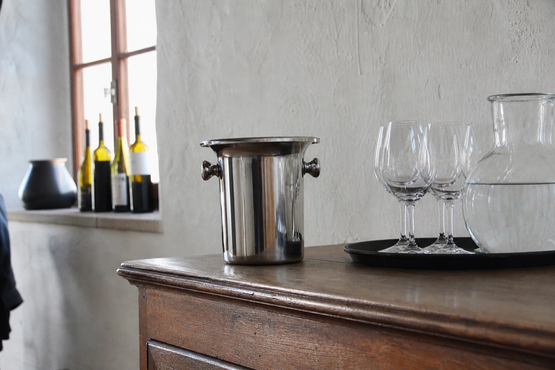 A stainless steel spittoon beside tasting glasses and a water jug on a wooden estate counter in soft daylight