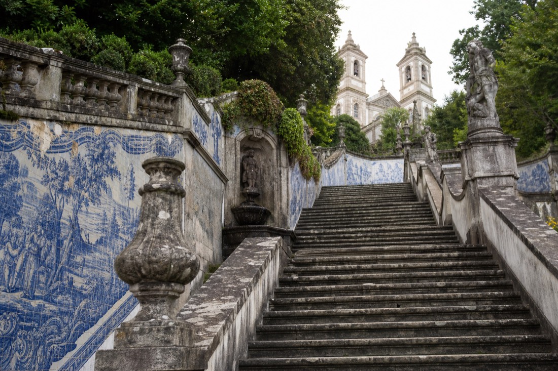 Wide baroque staircase with blue and white tiled walls, fountains and statues zigzagging up a wooded hillside